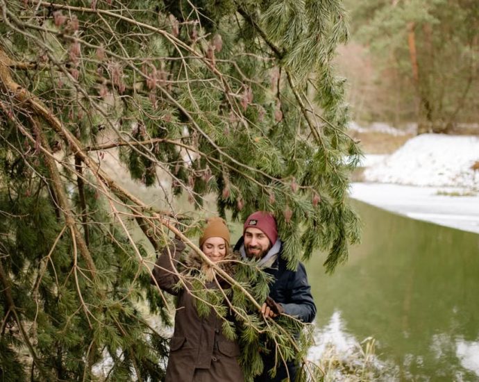 A happy couple exploring a snowy forest, smiling under pine trees by the river.