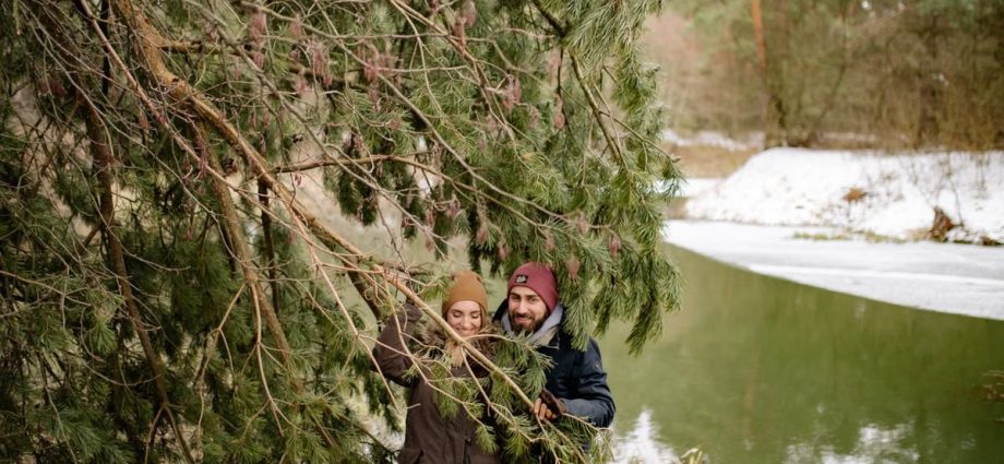 A happy couple exploring a snowy forest, smiling under pine trees by the river.