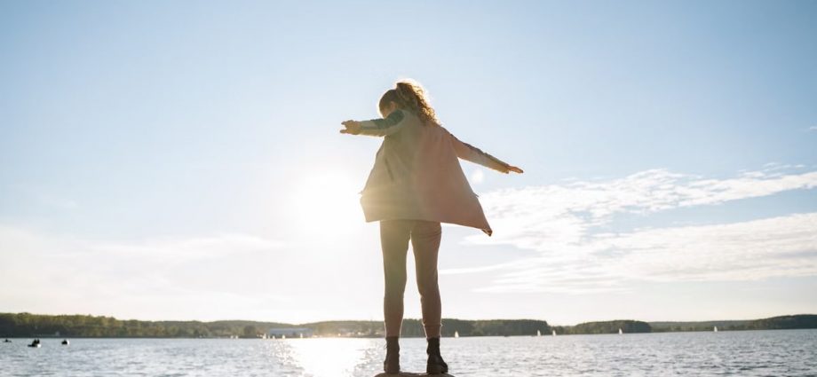 A woman stands on rocks, arms outstretched, enjoying a sunny day by a serene lake.